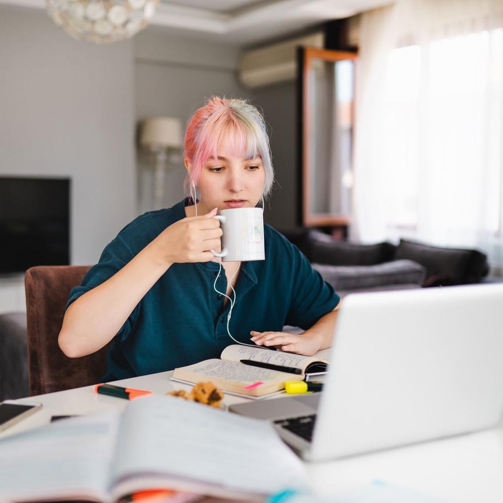 Eine junge Frau mit bunten Haaren sitzt an ihrem Laptop und trinkt aus einer Tasse.